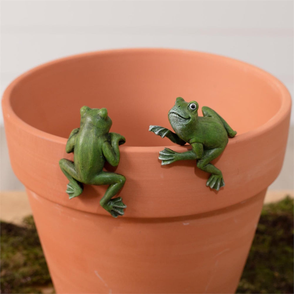 Two small green frog figurines climbing the rim of a terracotta flower pot.