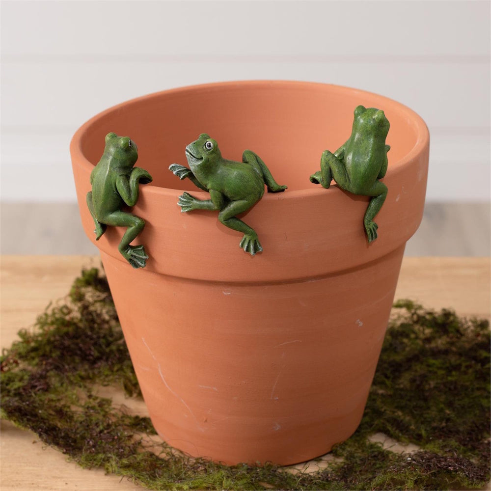 Three small green frog figurines climbing onto the rim of a terracotta plant pot.