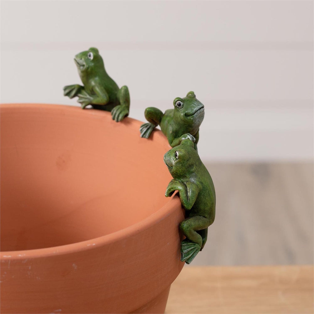 Three small green frog figurines climbing and perched on the rim of a terracotta flower pot.