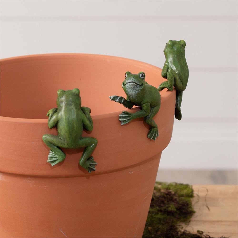 Three small green frog figurines climbing on the rim of a terracotta plant pot.