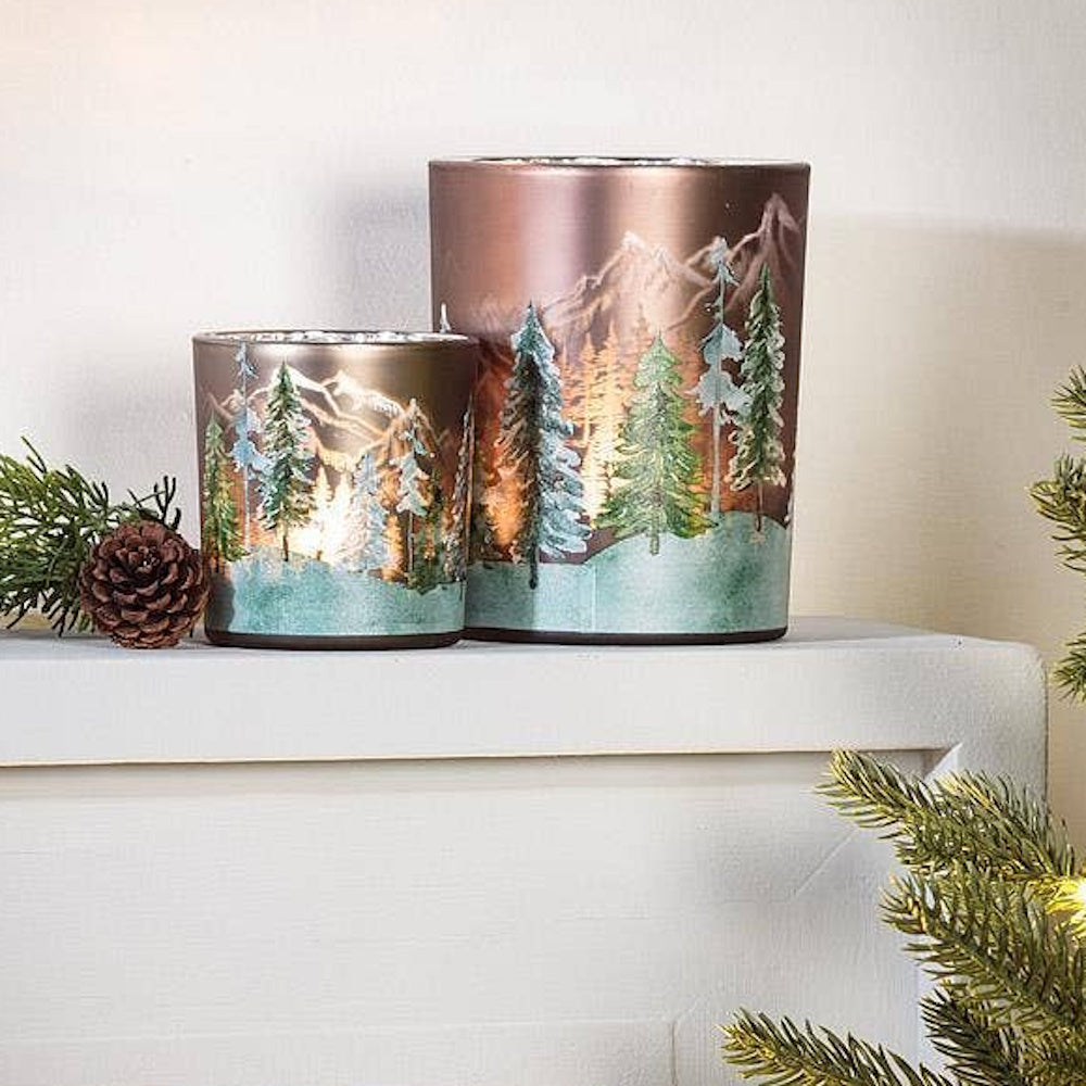 Two glowing glass votive candle holders featuring snowy pine trees and mountain landscapes.