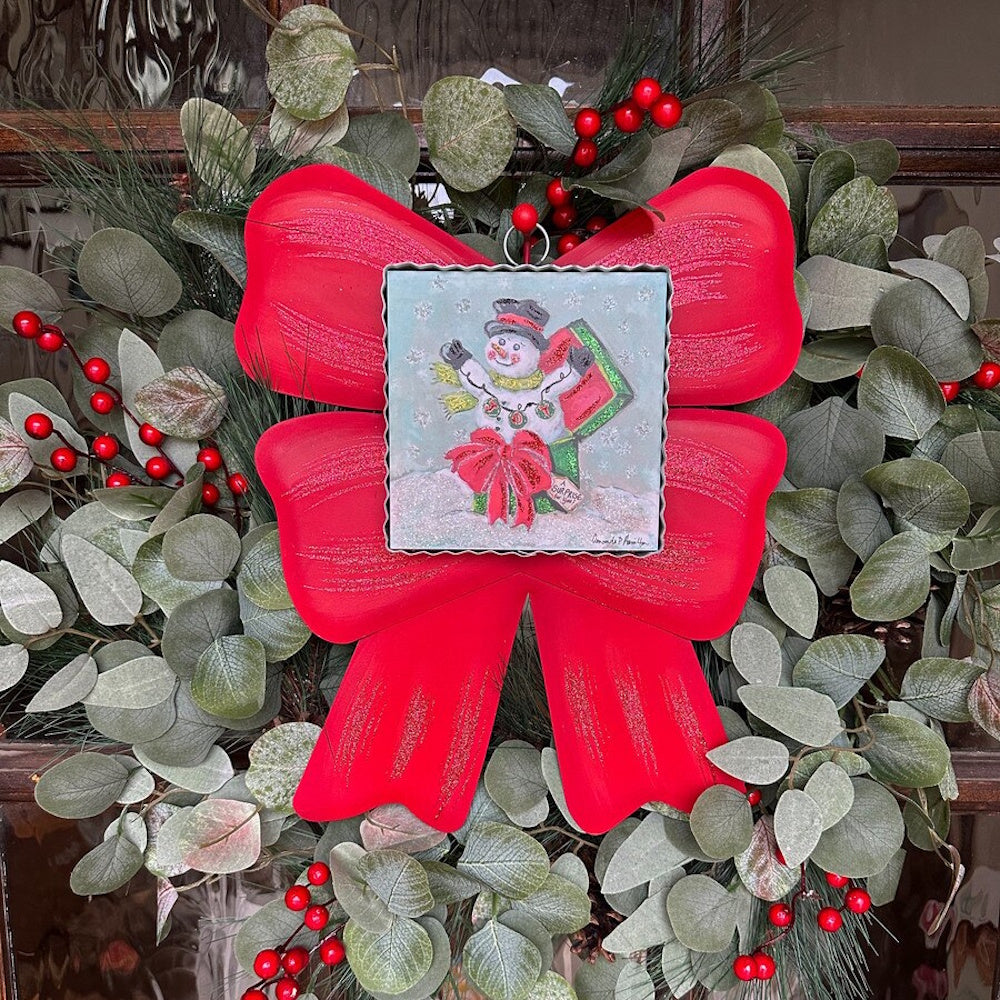 Front view of a square metal snowman art piece displayed on a large red bow and green holiday wreath.