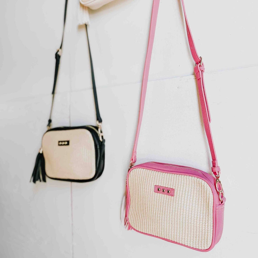 Two woven crossbody bags, one pink and one black and cream, hanging against a white wall.