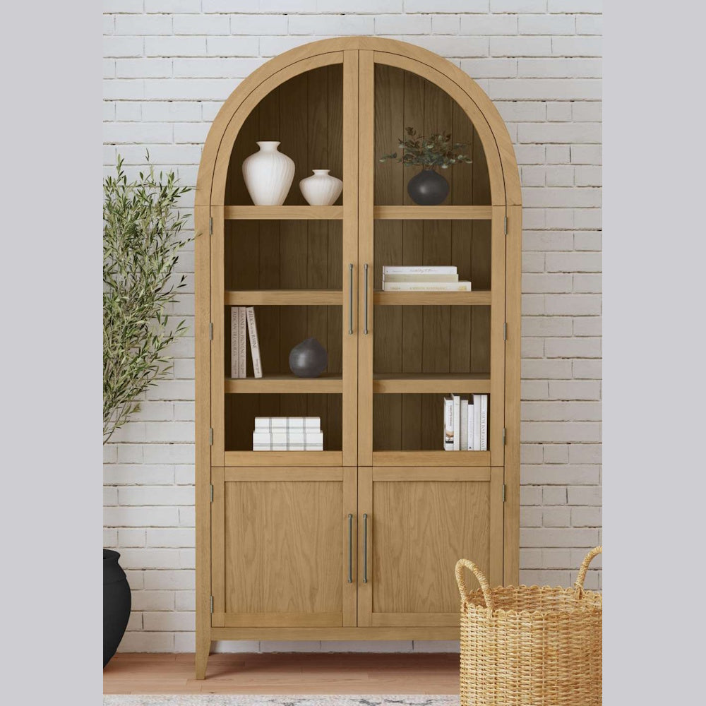 Arched light oak display cabinet with glass doors and shelving, styled with vases and books against a white brick wall.