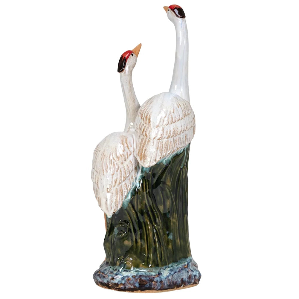 Two glazed ceramic cranes with white feathers and red crowns stand together on a green and blue base.
