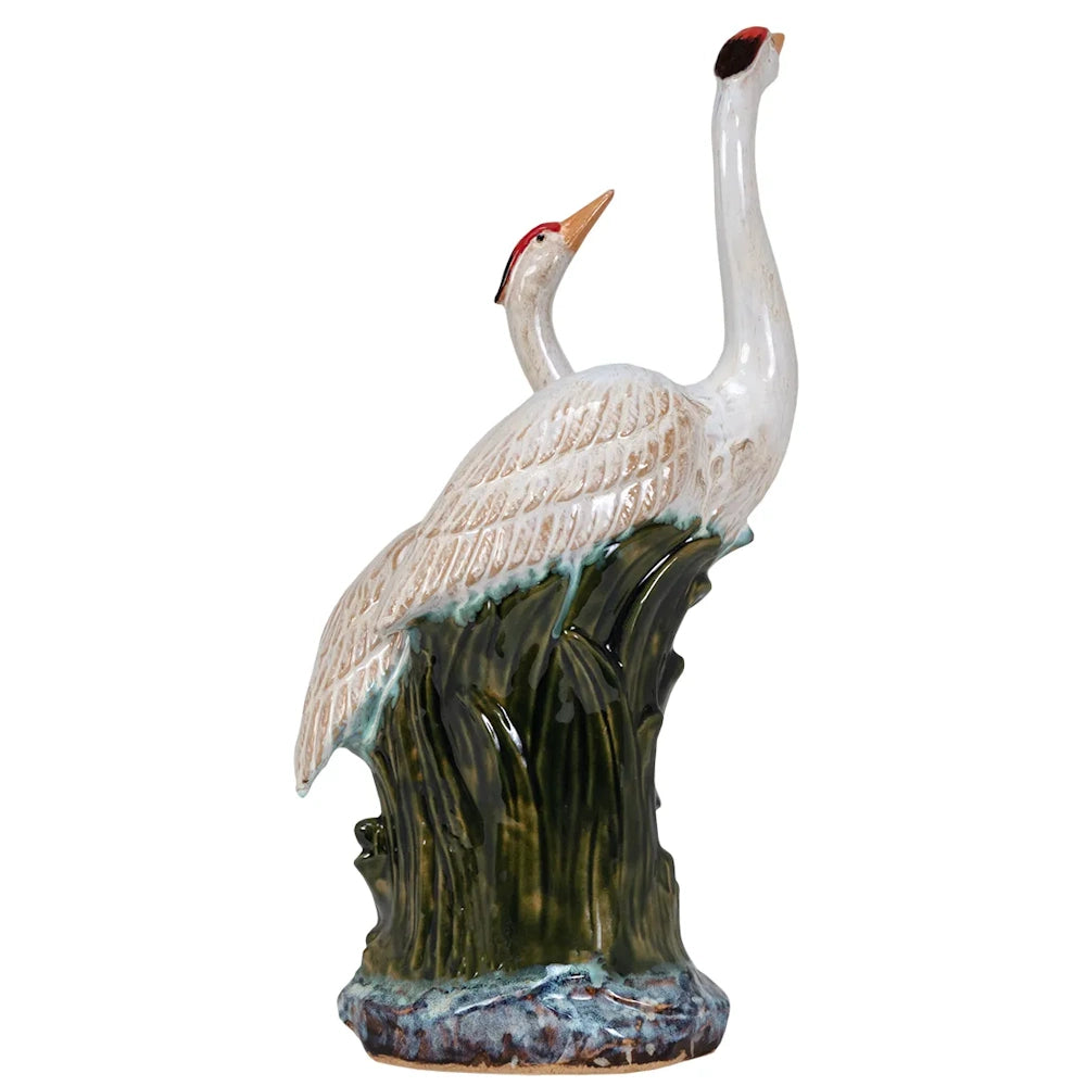 Side view of a ceramic figurine depicting two white herons with red crests standing on a glossy green base.
