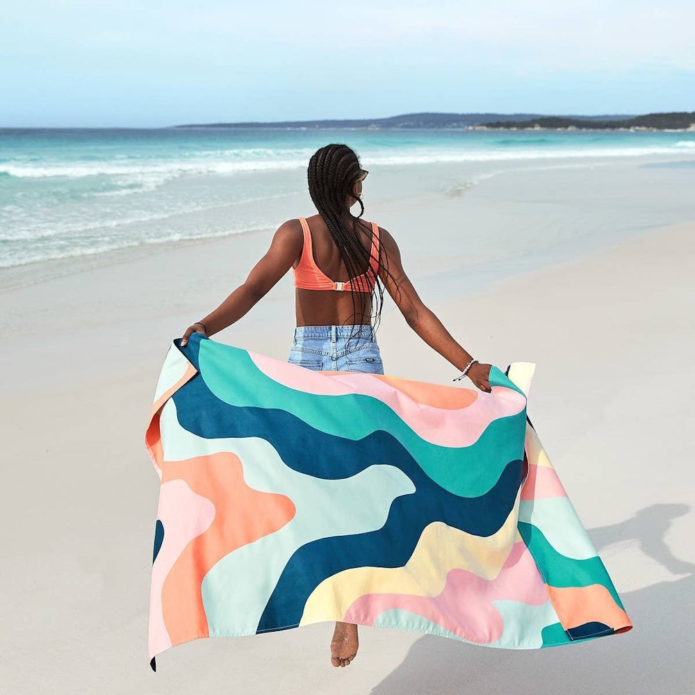 Woman walking on a beach holding a large, quick-dry towel with a colorful, wavy abstract pattern.