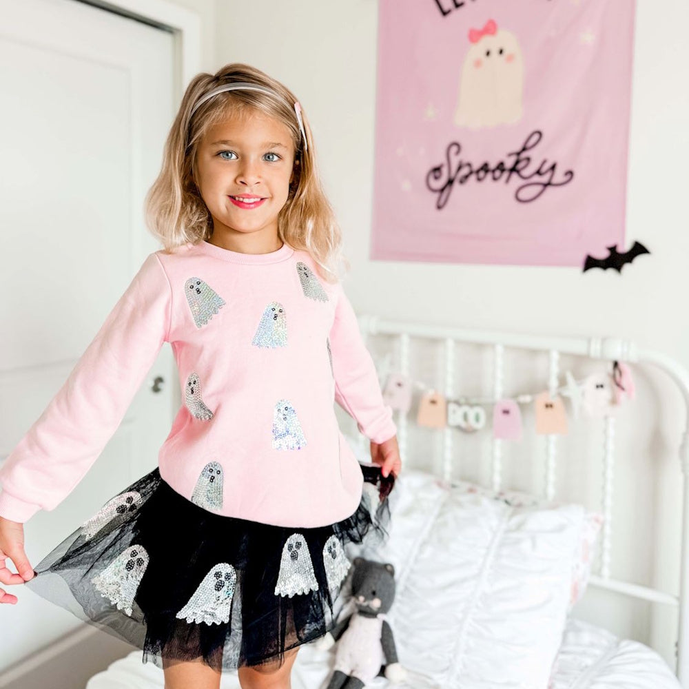 A young girl smiles in a pink ghost sweatshirt and a black tutu skirt while standing on a bed.