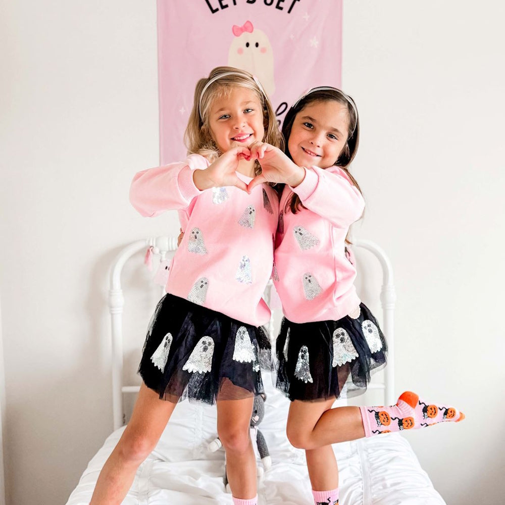 Two girls wearing pink sequin ghost sweatshirts and black skirts make a heart shape with their hands.