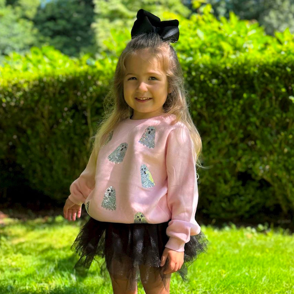 A young girl wears a pink ghost sweatshirt and a black tutu skirt while posing outside in front of green bushes.