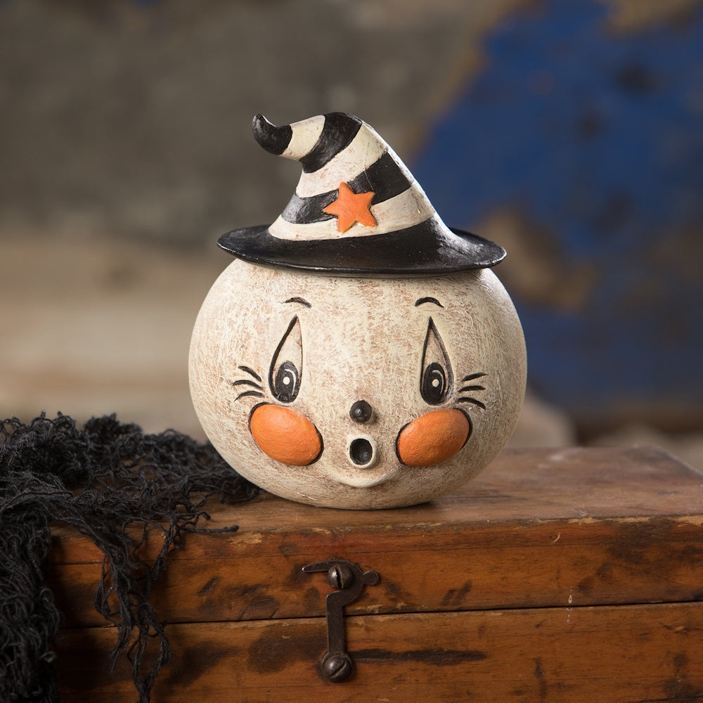 A vintage round ghost figurine with rosy cheeks and a striped witch hat sitting on a wooden chest.
