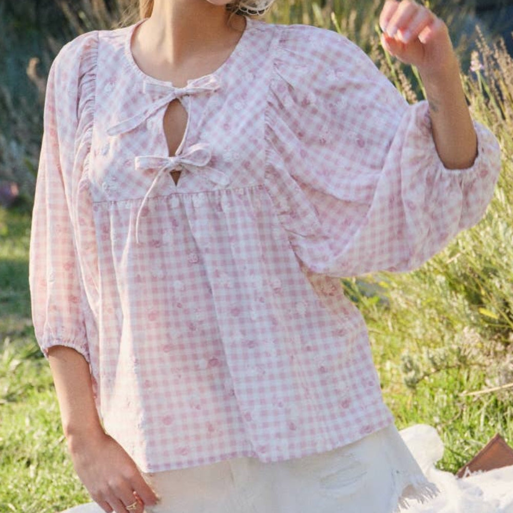 A woman in a grassy field wearing a pink gingham babydoll top with puff sleeves and tie details at the neckline.