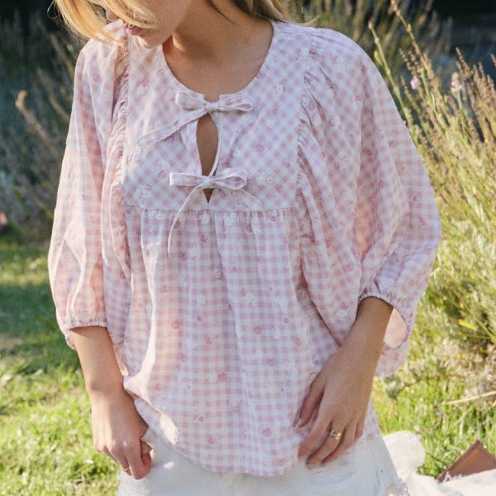 Pink and white gingham blouse with floral details, puff sleeves, and double bow ties on a woman outdoors.