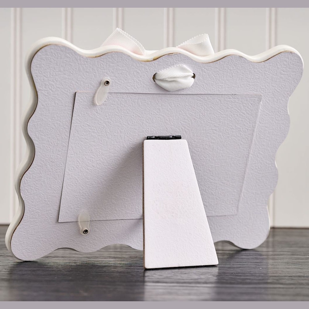 Back view of a scalloped picture frame showing the easel stand, turn buttons, and a white decorative bow.