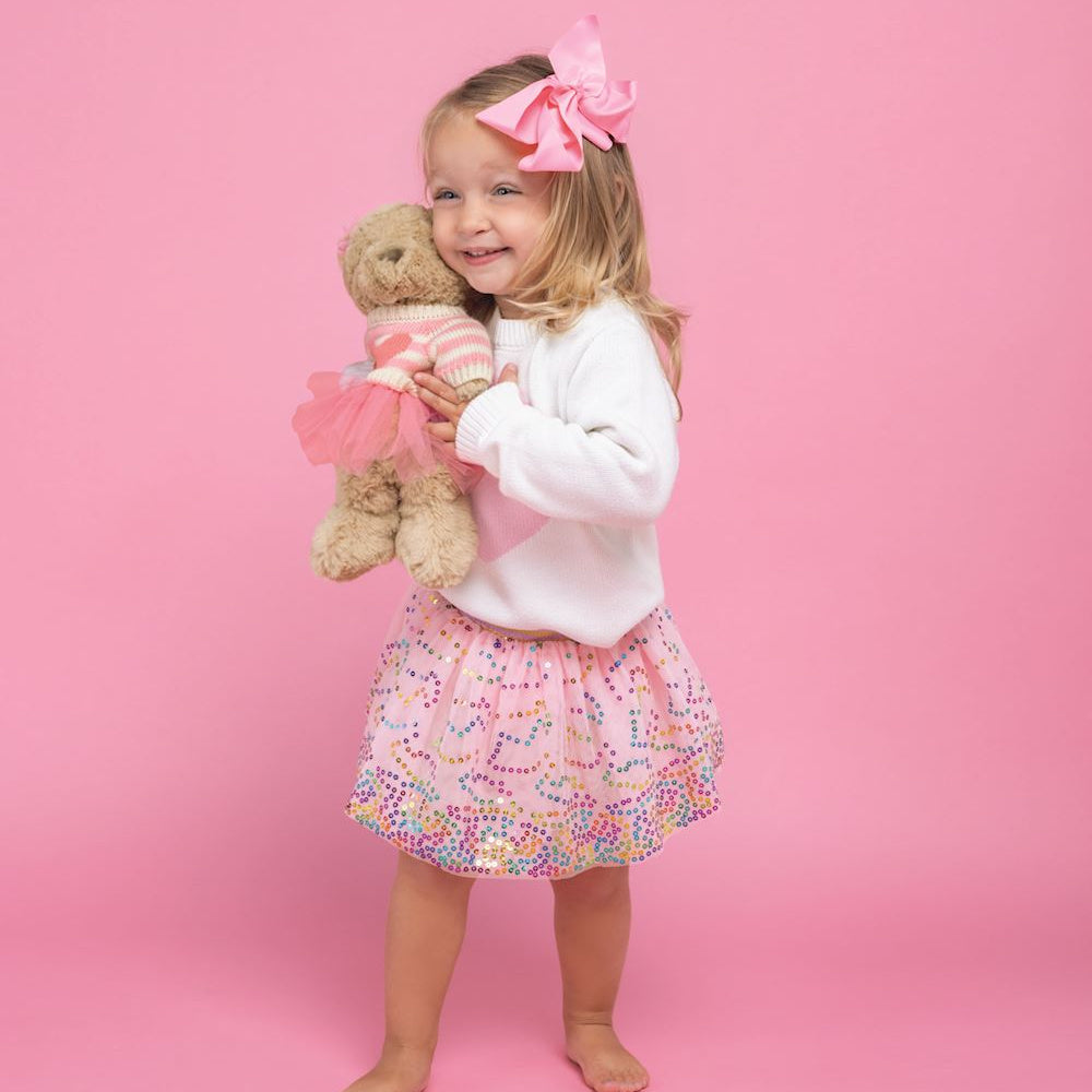 Little girl in white sweater and pink sequin tutu holding a teddy bear in a matching pink outfit on a pink background.