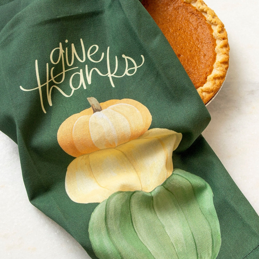 Green kitchen towel with "give thanks" text and stacked pumpkins next to a pumpkin pie.