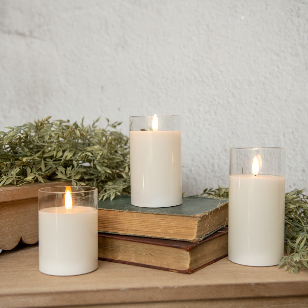 Three glowing white flameless pillar candles in glass holders displayed with vintage books and greenery.