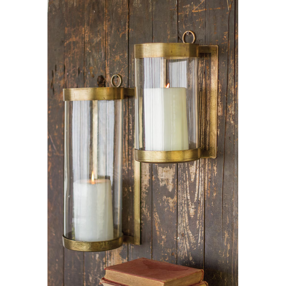 Pair of wall-mounted antique brass hurricane candle sconces with glass cylinders on wooden wall.