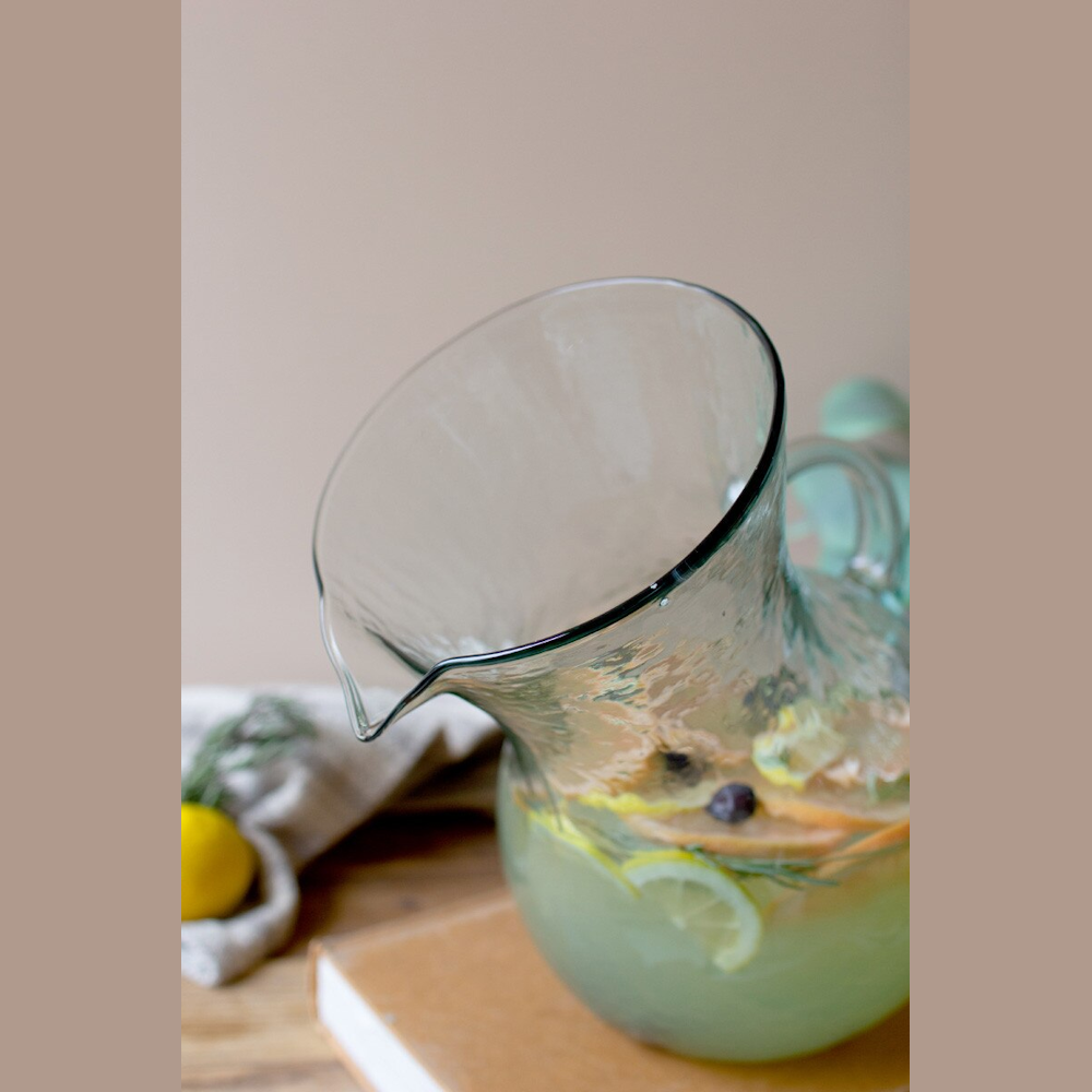 Tilted glass pitcher of citrus-infused water with lemon, grapefruit slices, and blueberries on a wooden table.