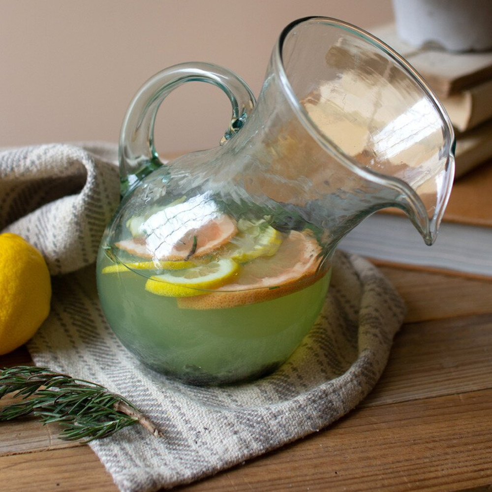 Tilted hand-blown glass pitcher filled with citrus infused water and rosemary on a rustic wooden table.