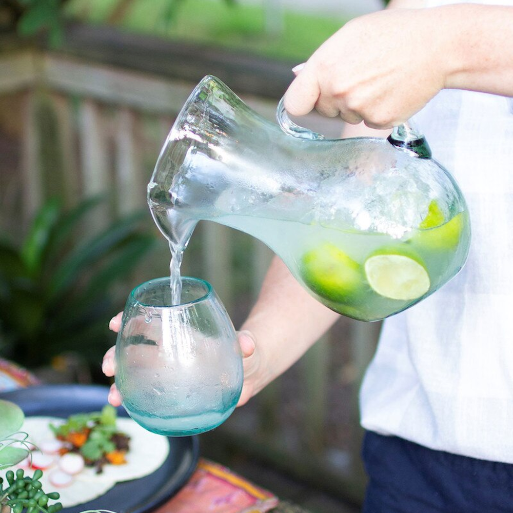 A person pouring fresh lime-infused water from a clear glass pitcher into a matching blue-tinted glass outdoors.
