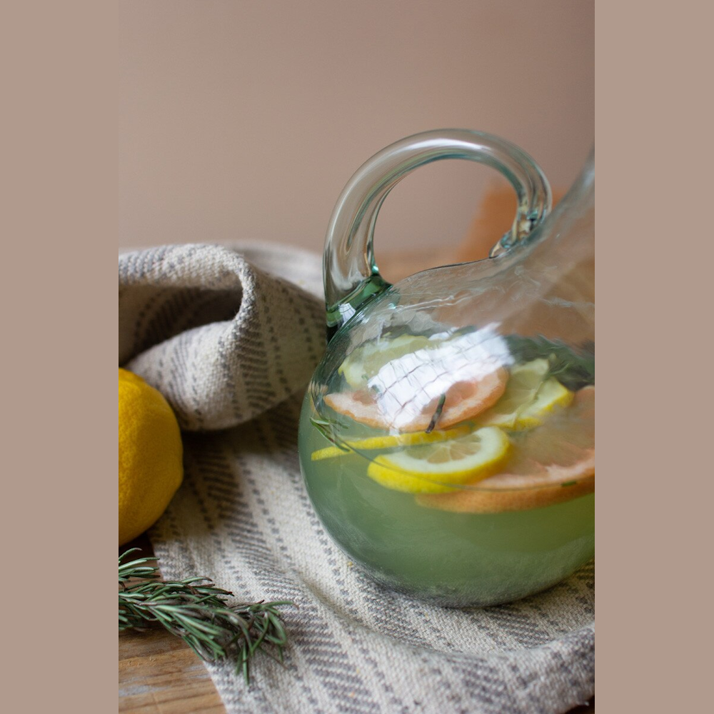 Infused citrus water in a round glass pitcher with fresh rosemary and lemon on a rustic linen cloth.