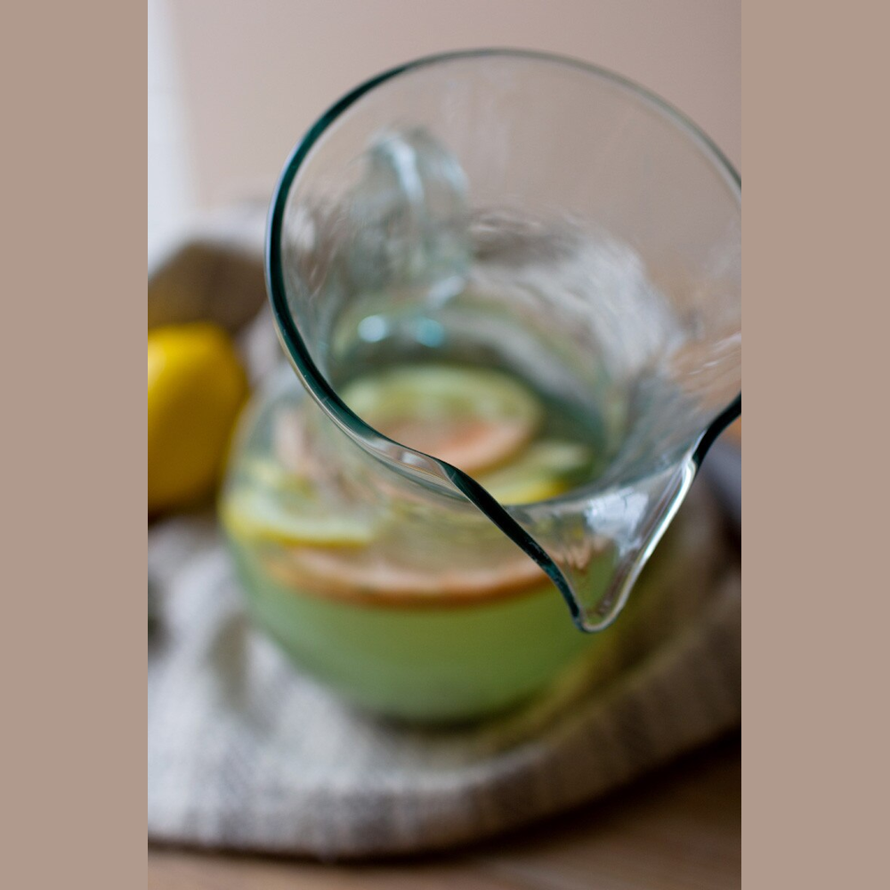 Tilted clear glass pitcher filled with infused green liquid and citrus slices on a rustic linen cloth.