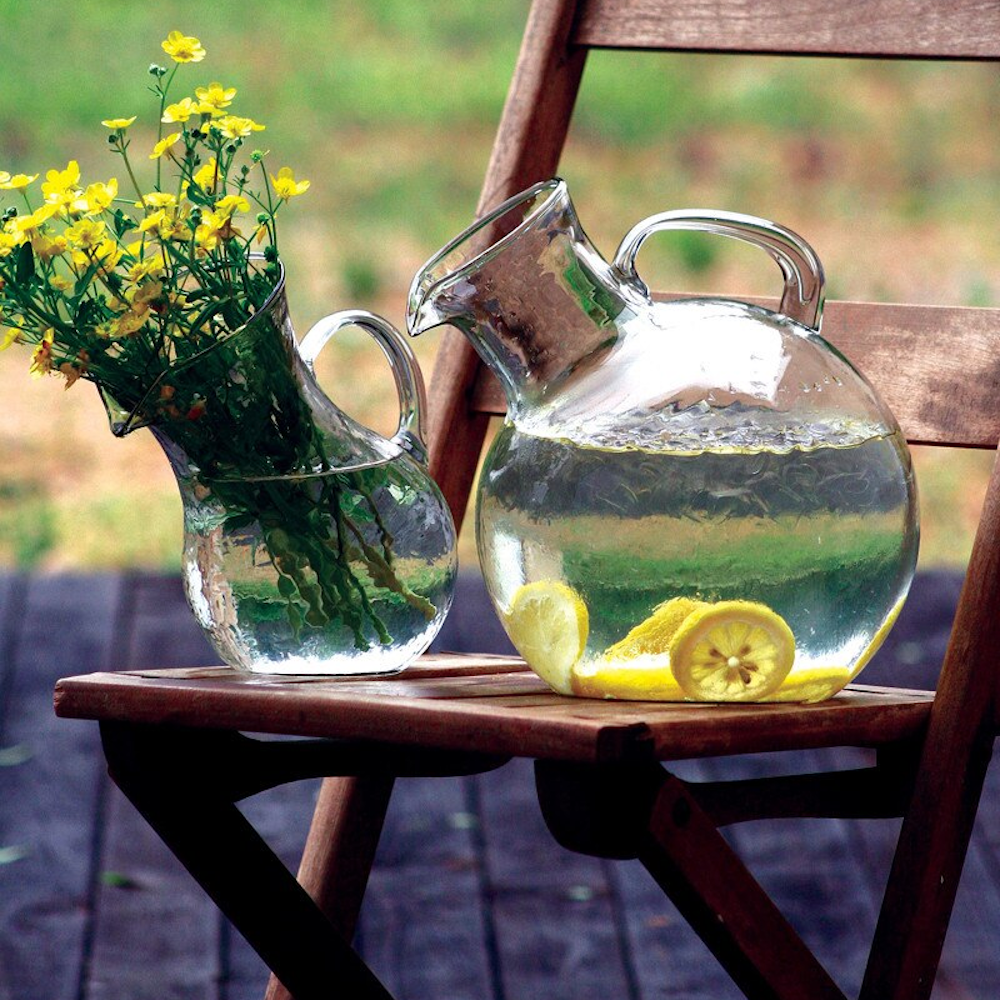 Tilted hand-blown glass pitchers with yellow flowers and lemon-infused water on a rustic wooden folding chair outdoors.