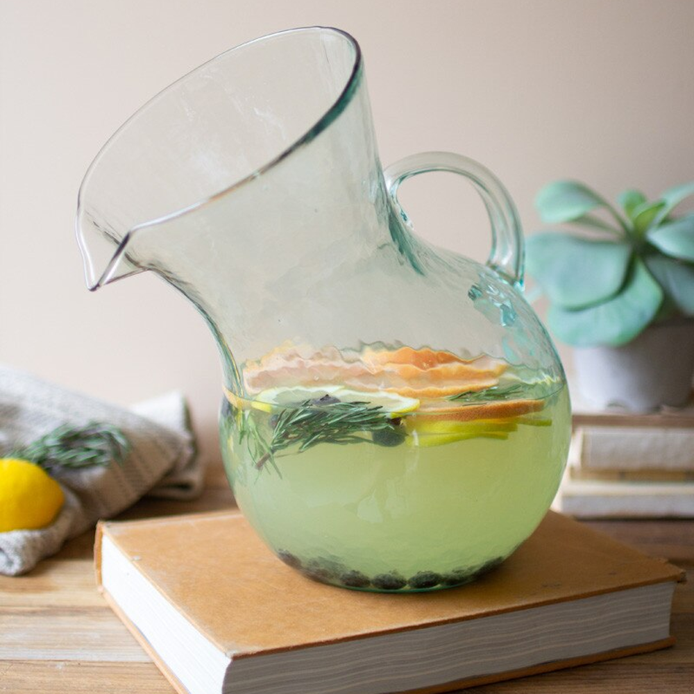 Tilted hand-blown glass pitcher with citrus infused water and rosemary sitting on a vintage book.