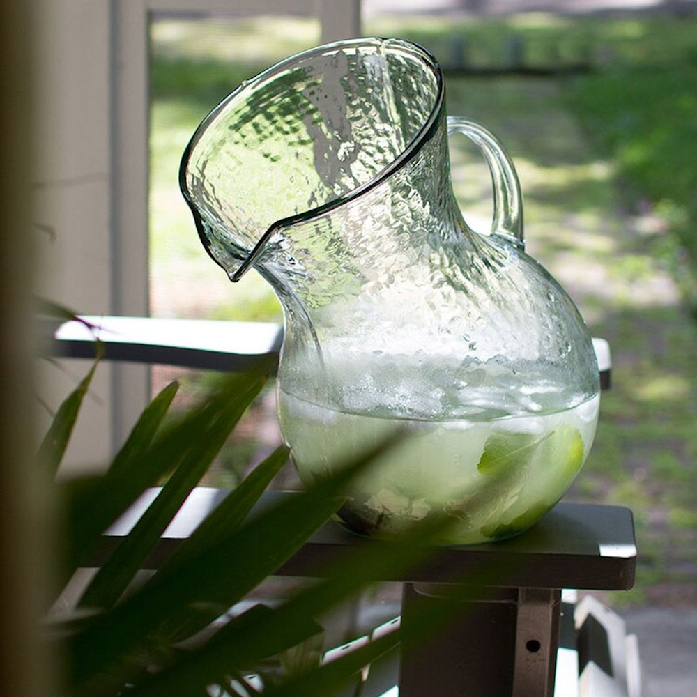 Tilted hammered glass pitcher filled with ice and lime water sitting on a porch railing with garden background.