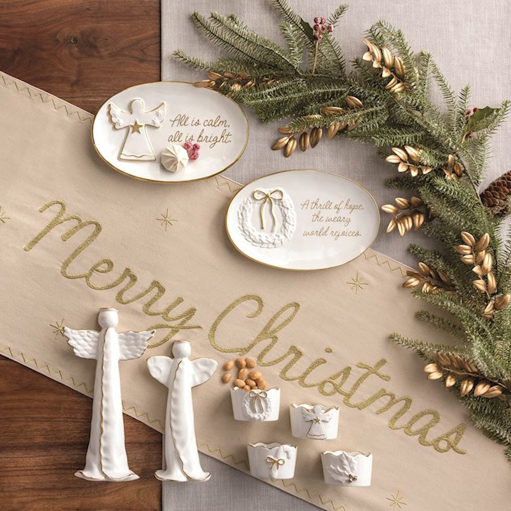 White ceramic angel statues, trays, and dishes with gold trim on a Merry Christmas table runner.