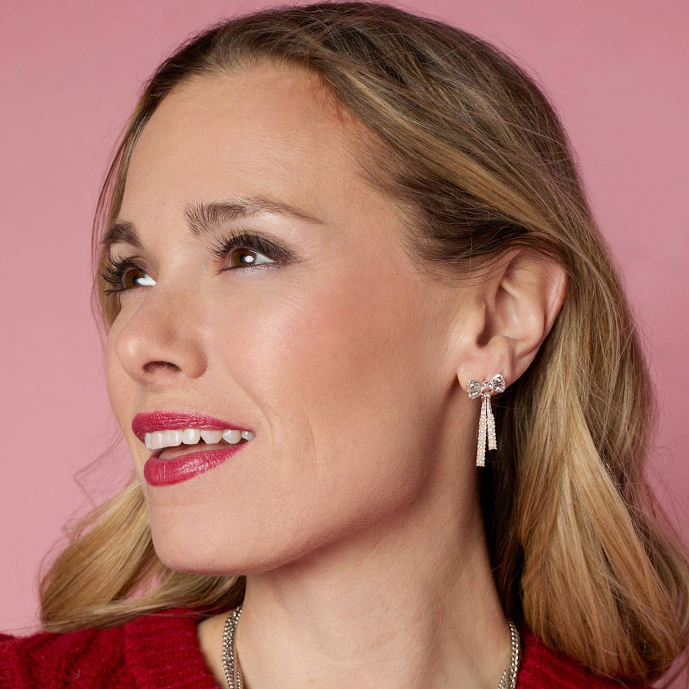 Woman smiling in profile, wearing a gold and diamond bow-shaped dangle earring against a pink background.