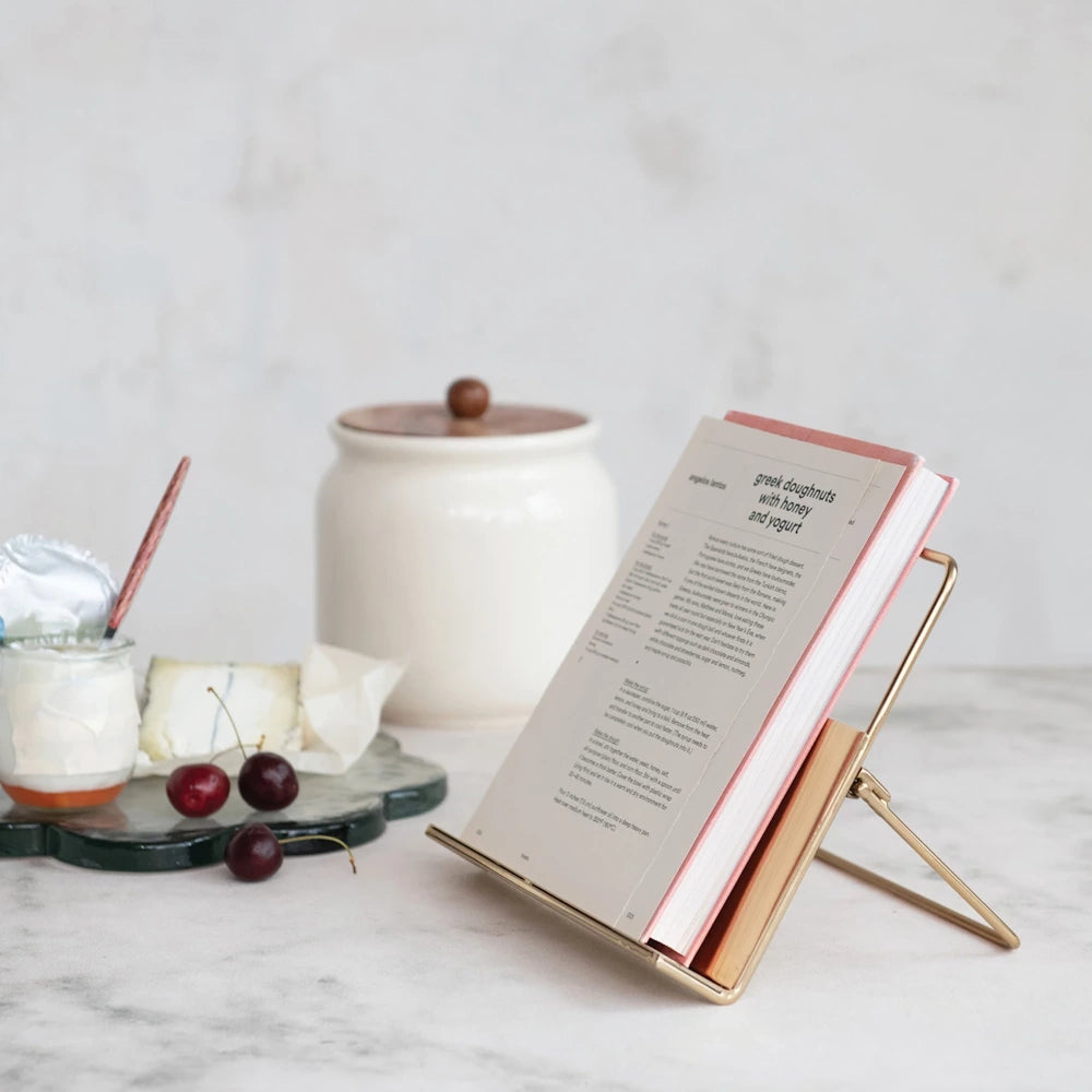 Elegant gold wire cookbook stand holding an open recipe book on a marble kitchen counter with rustic decor.