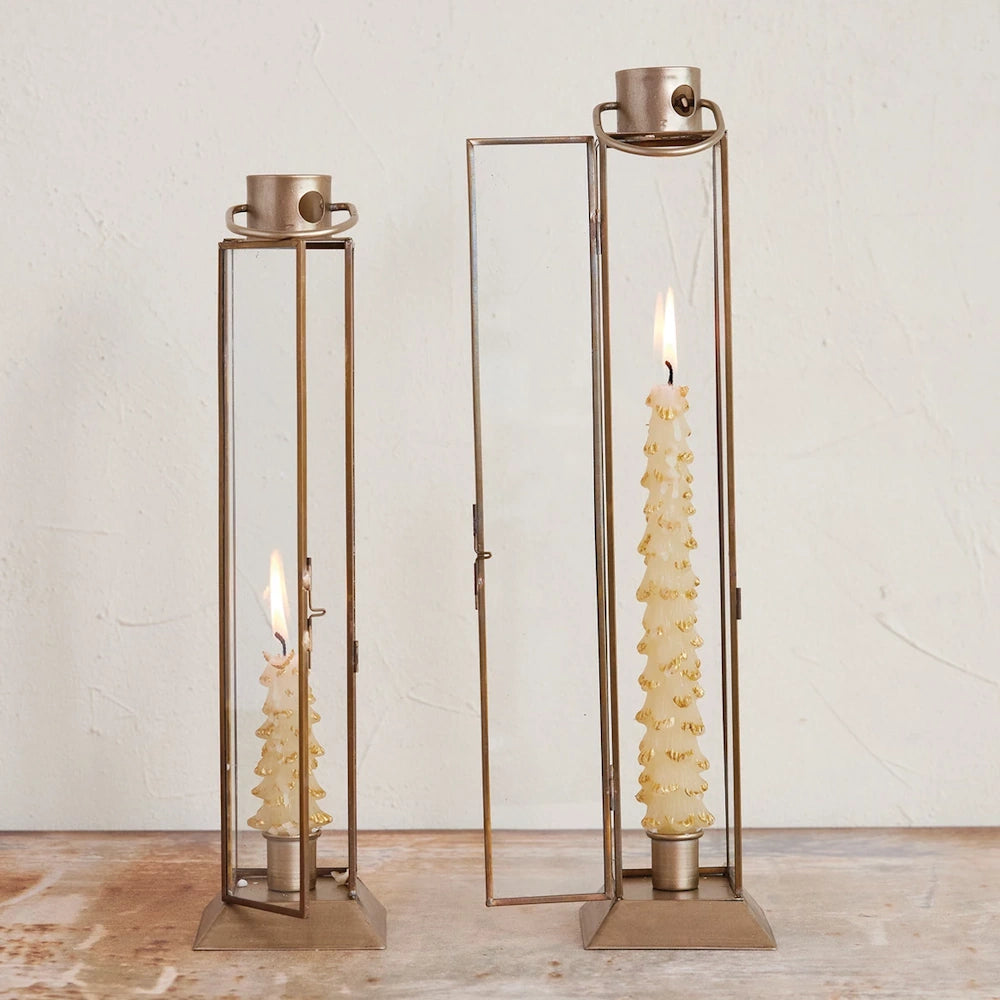 Gold-finish metal and glass lantern taper holders with lit tree-shaped candles against a white textured wall.