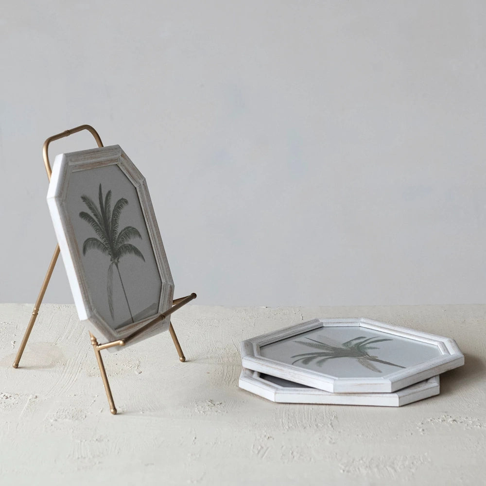 Gold-finish metal tabletop easel displaying a palm tree art print in a white octagonal frame on a textured surface.