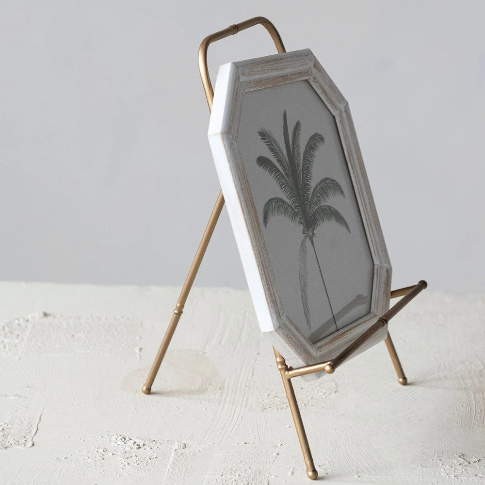Gold-finish metal easel holding an octagonal white wooden frame with a palm tree sketch on a textured white surface.