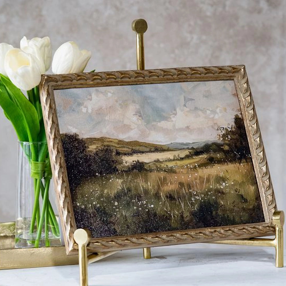 Framed vintage landscape oil painting of a meadow in a gold ornate frame on a stand next to white tulips.