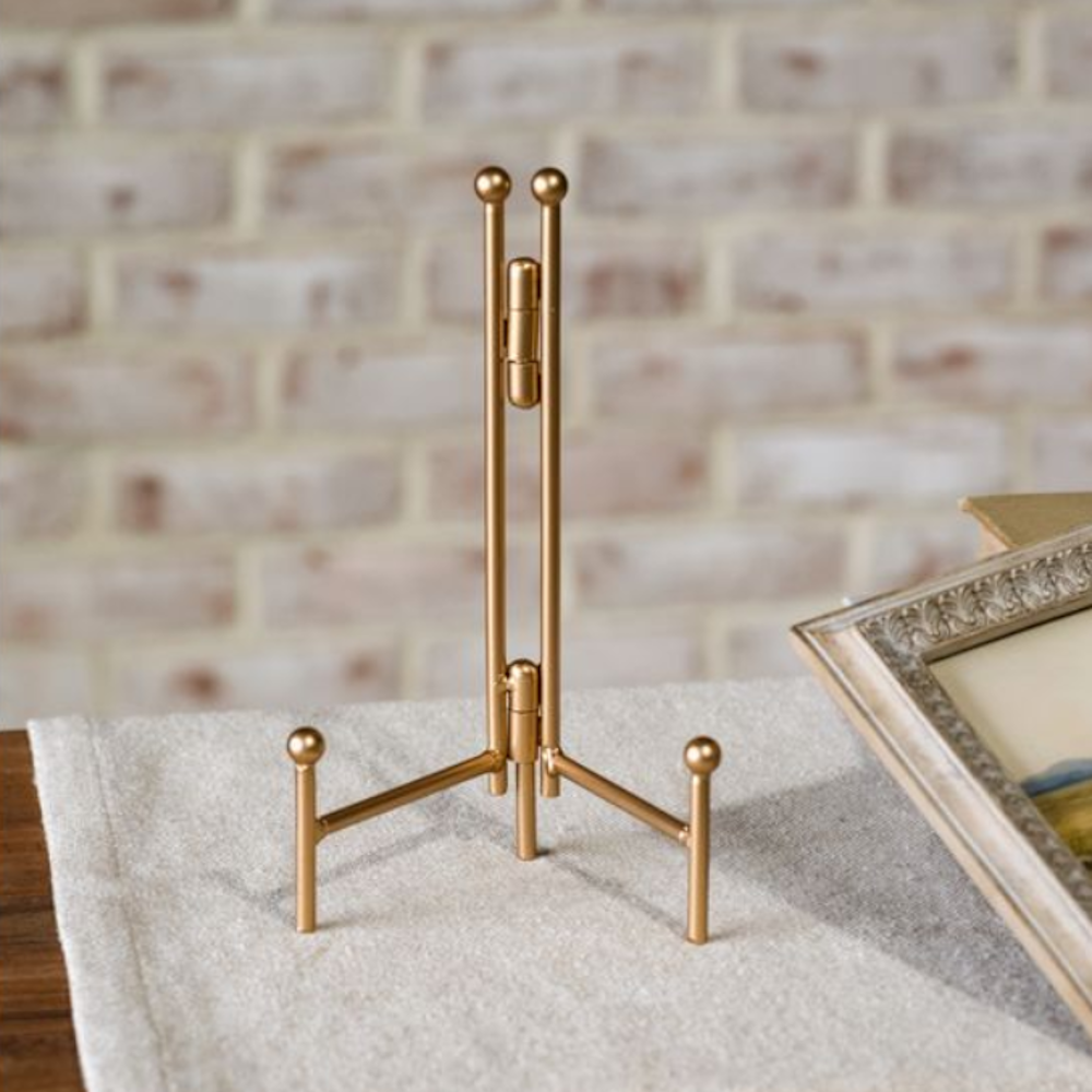 Gold metal tabletop display easel standing on a tablecloth against a light brick background.