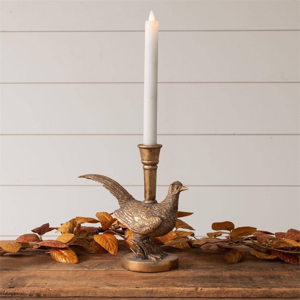 A gold-tone pheasant candleholder with a tall white taper candle on a wooden table decorated with autumn leaves.
