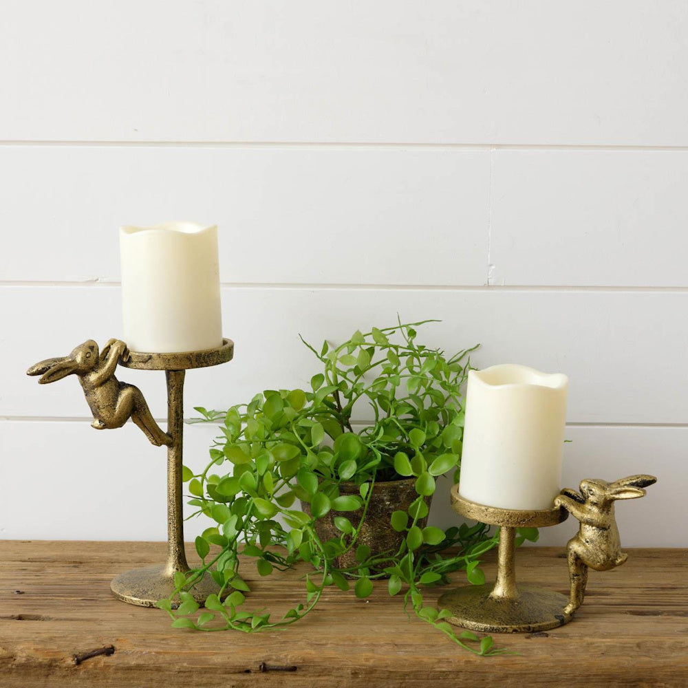 Two gold rabbit pillar candle holders in varying heights with white candles on a rustic wood surface with green foliage.