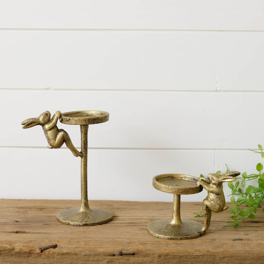 Two gold rabbit pillar candle holders in different heights on a rustic wood surface against a white shiplap wall.