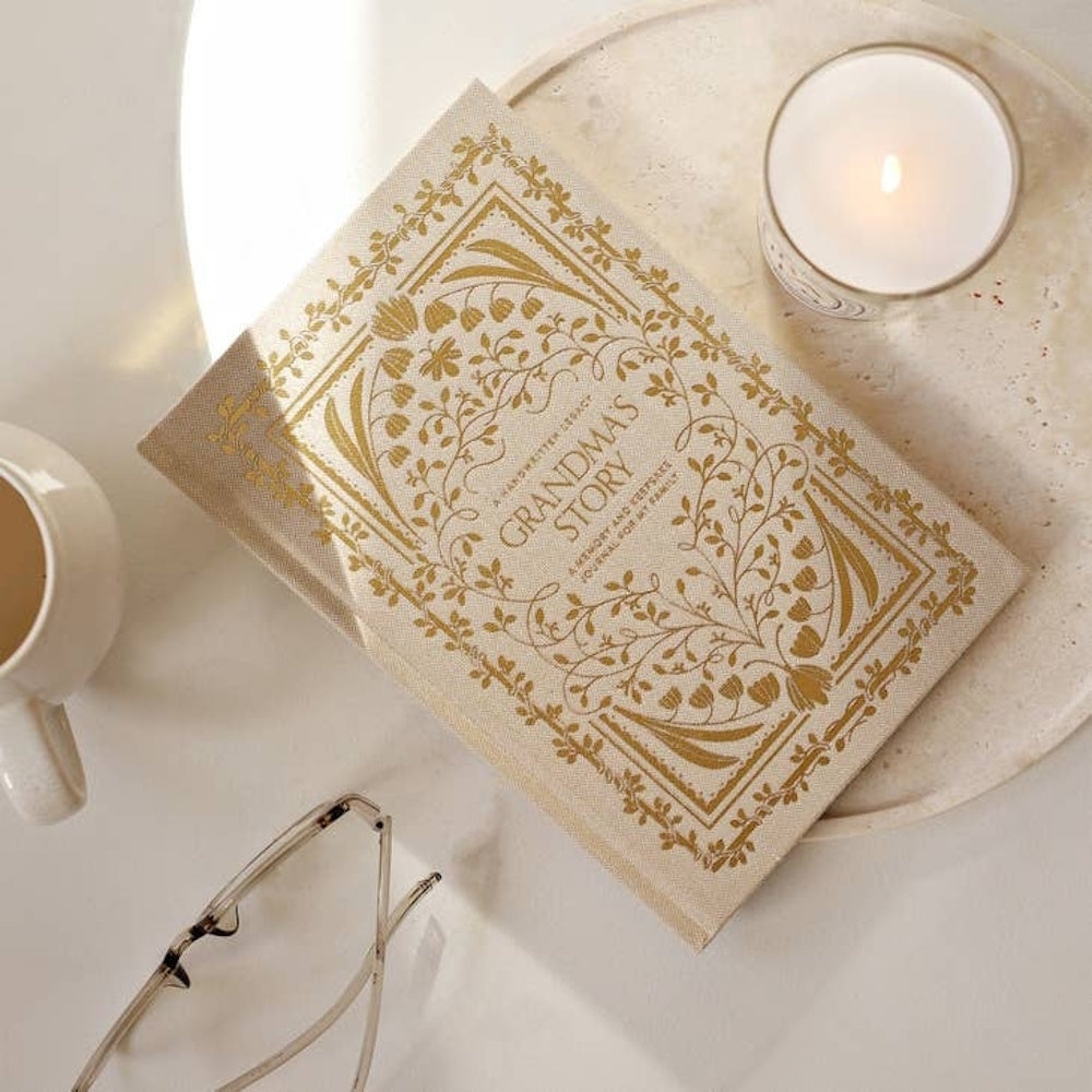 Grandma's Story keepsake journal with gold floral patterns on a desk next to a lit candle, coffee cup, and glasses.