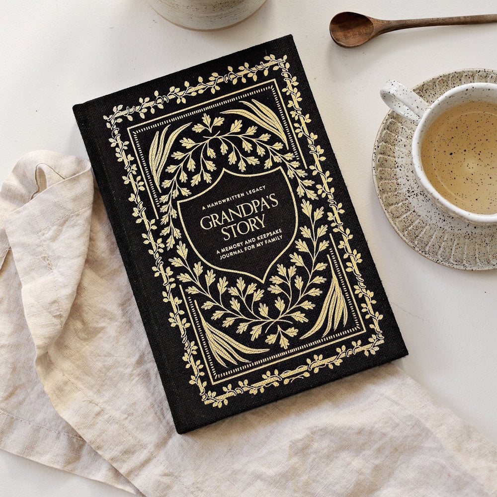 Black "Grandpa's Story" keepsake journal with gold leaf patterns on a white table next to a teacup and linen napkin.