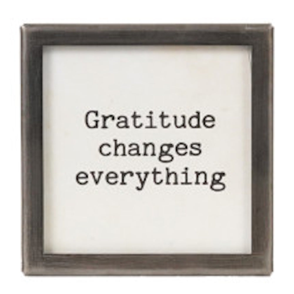 A square silver-framed sign with the text "Gratitude changes everything" in black typewriter font.