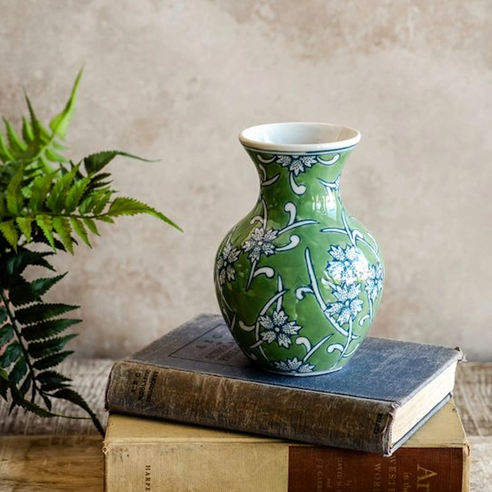 A green patterned porcelain vase sitting atop a stack of two old books, with a fern plant on the left.