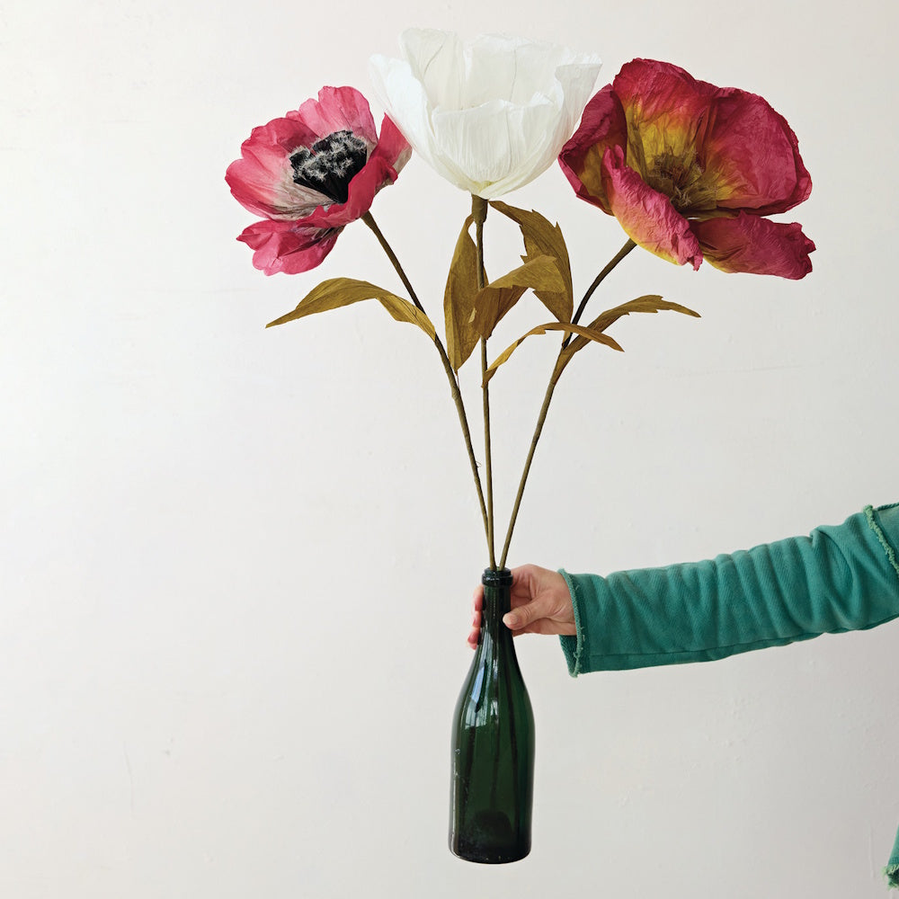 Hand holding large crepe paper flower picks in burgundy and white inside a green glass bottle vase.