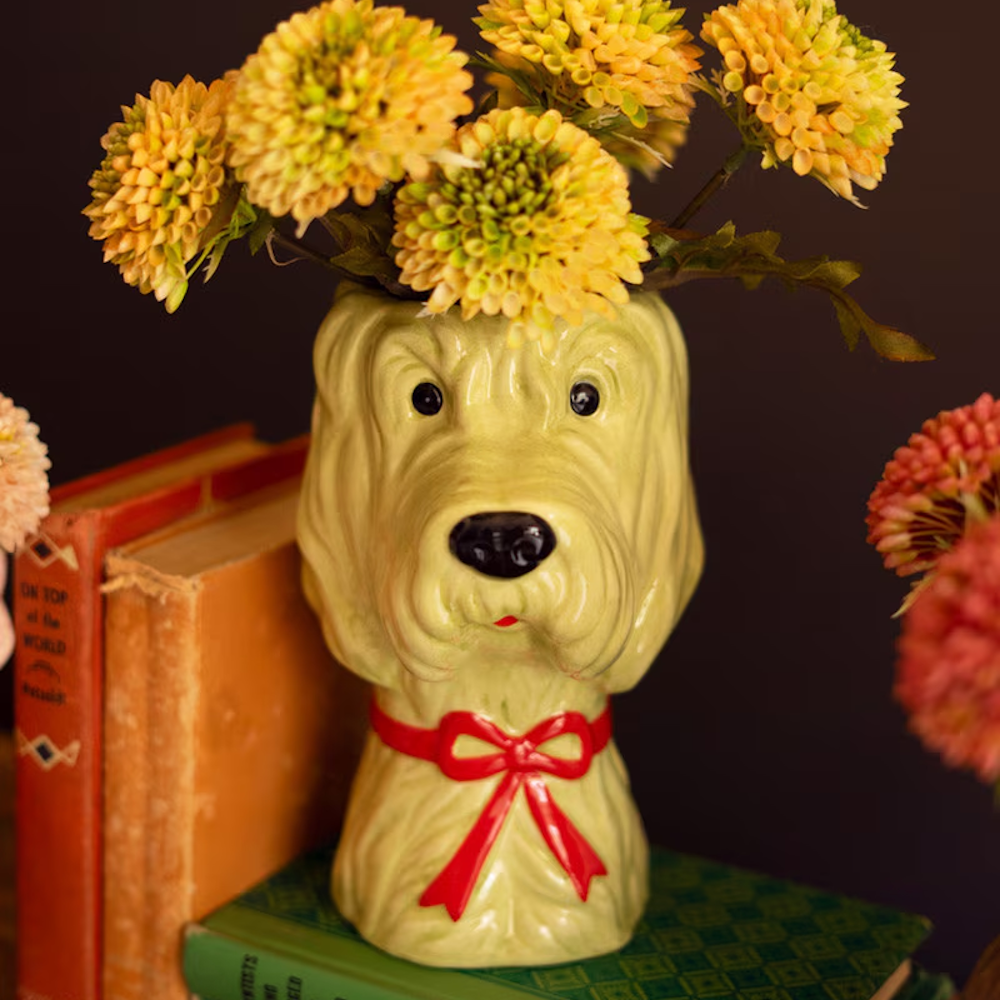 Green ceramic dog-head vase with a red bow and yellow flowers, sitting on vintage books against a dark background.