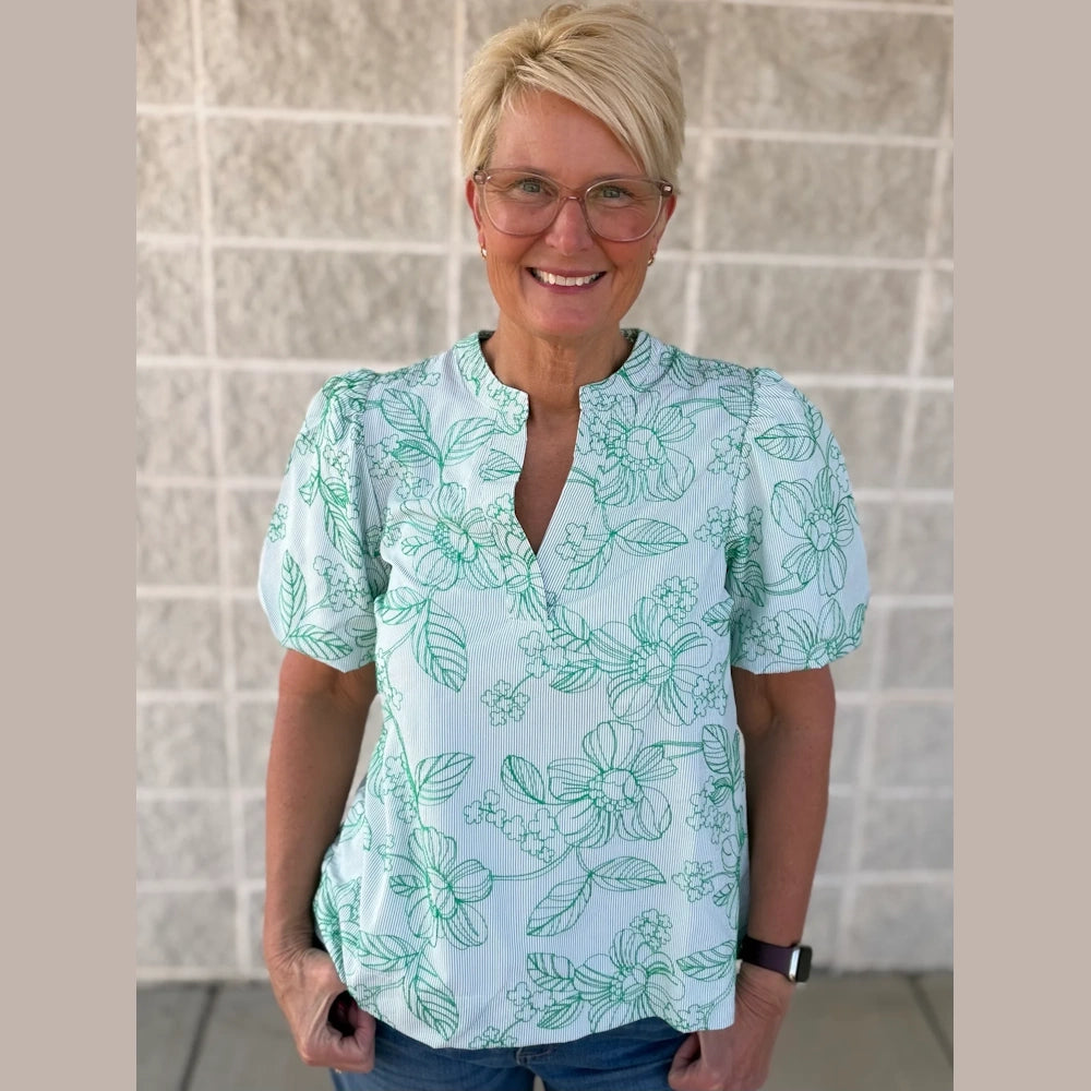 A woman with blonde short hair smiles in a green floral blouse against a textured white wall.