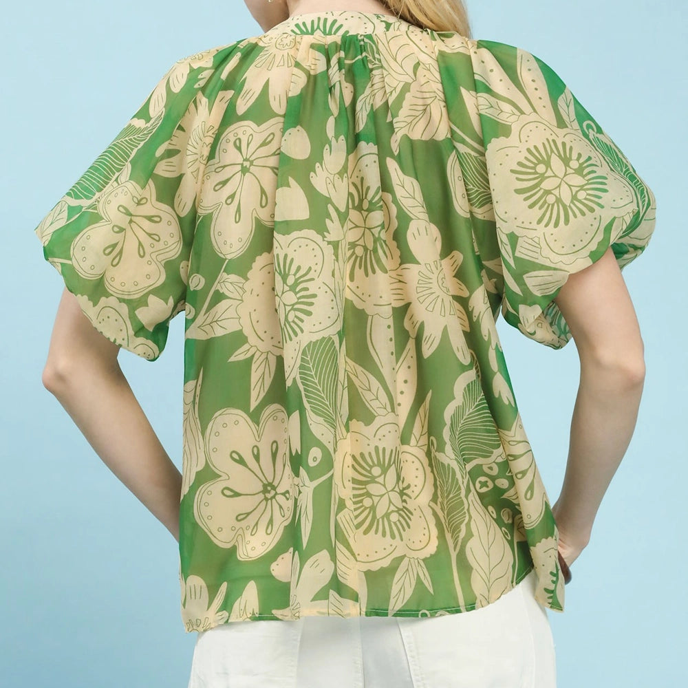 Back view of a woman wearing a green and cream floral puff-sleeve blouse with white pants against a blue background.