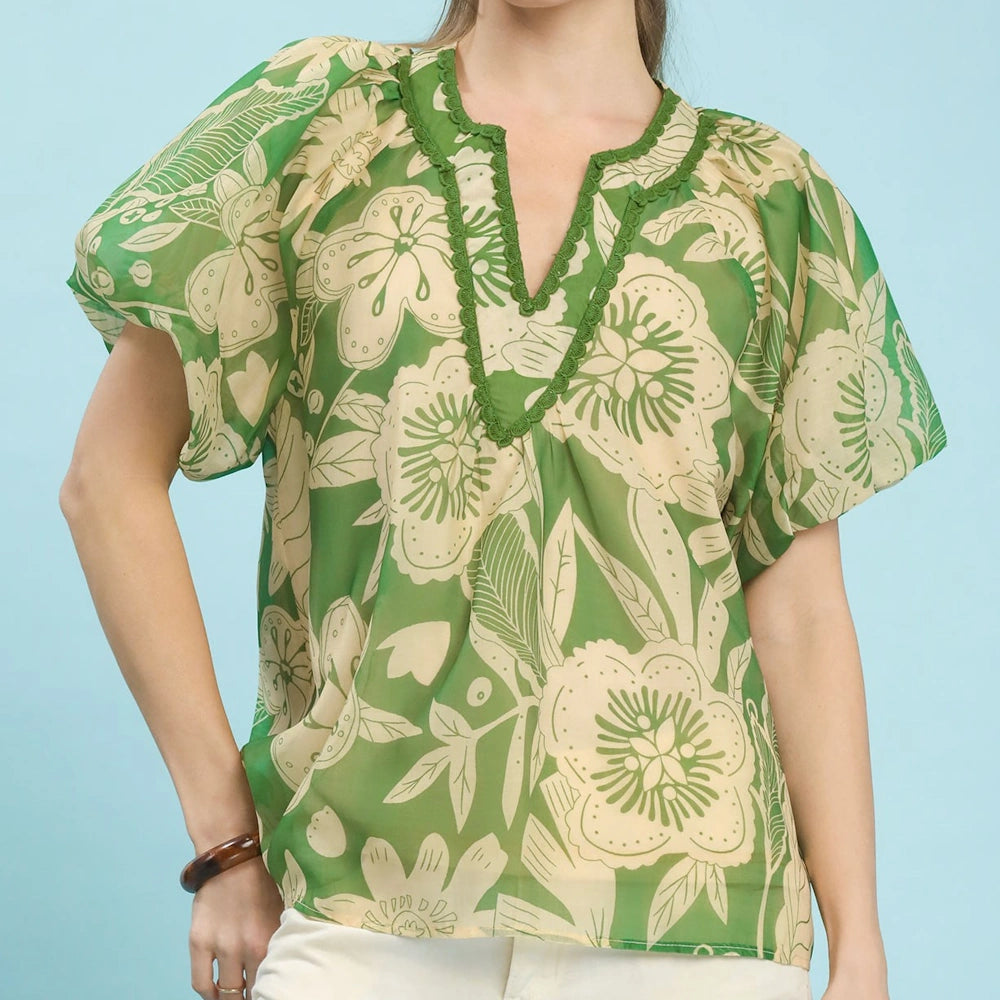 Woman wearing a green and cream floral v-neck blouse with puff sleeves and scalloped trim against a blue background.